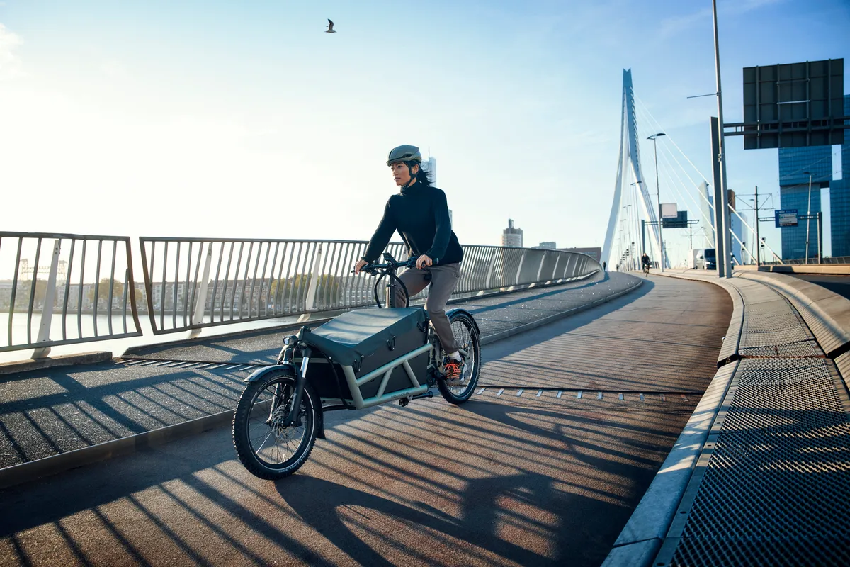 A lady riding a tundra grey matt Riese & Müller Load4 60 cargo bike on a cycle lane across a bridge.