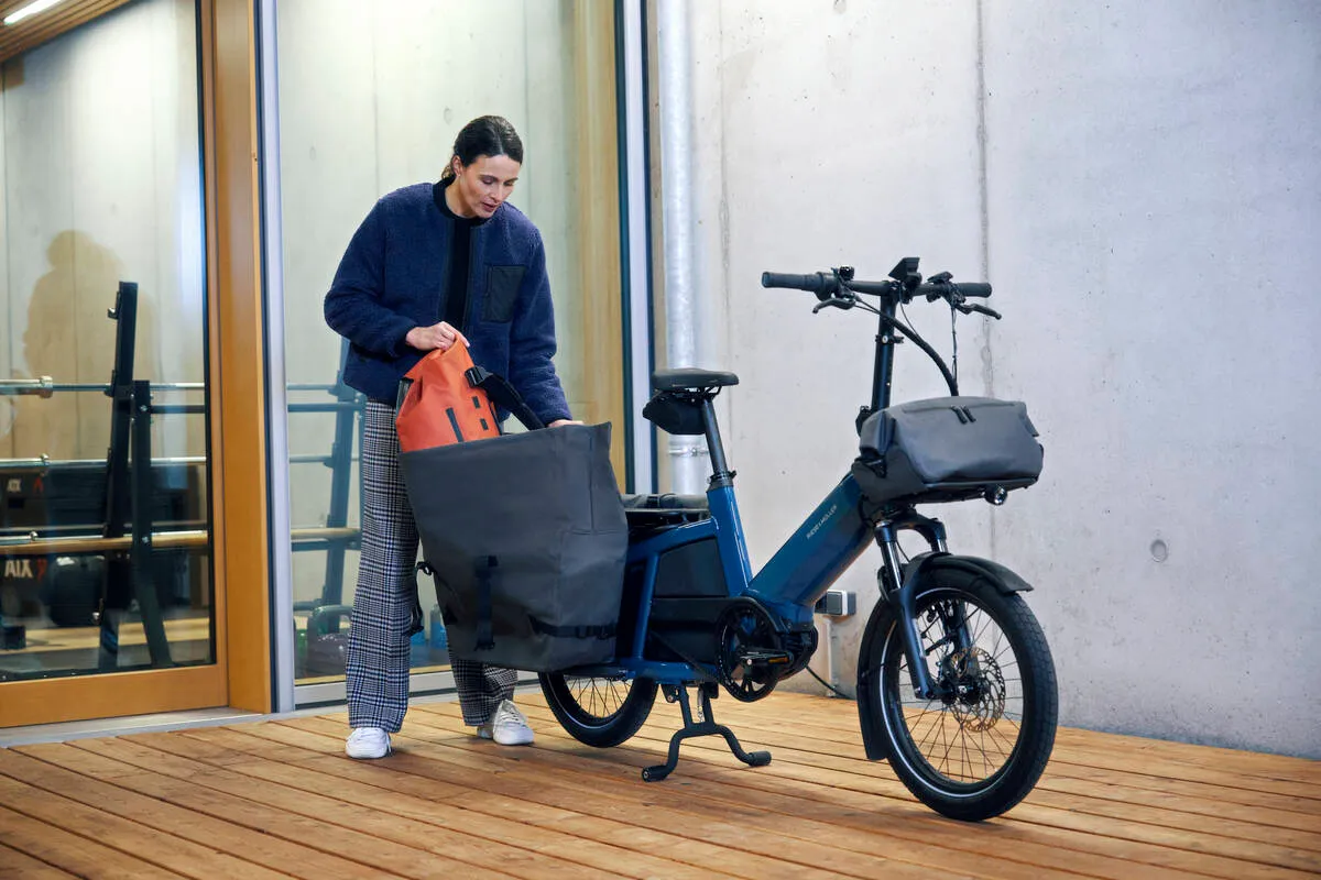 A cyclist loads bags onto a blue Riese & Müller Multitinker e-bike on a wooden deck outside a building.
