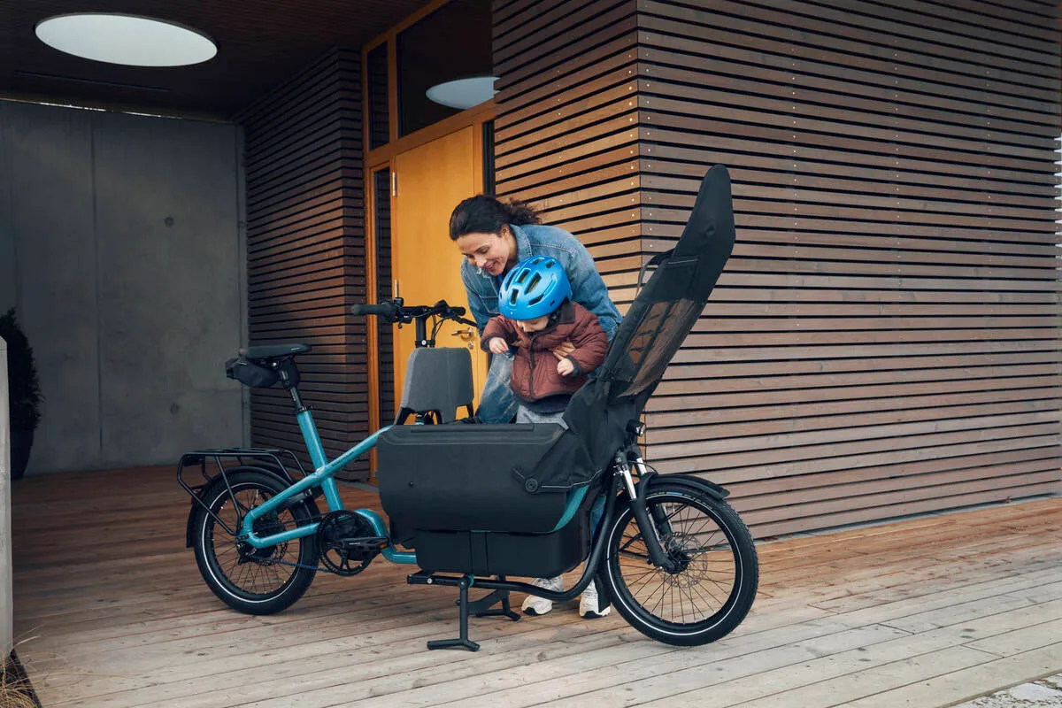 A child in blue helmet being secured in Riese & Müller Carrie e-cargo bike with aqua frame outside modern home