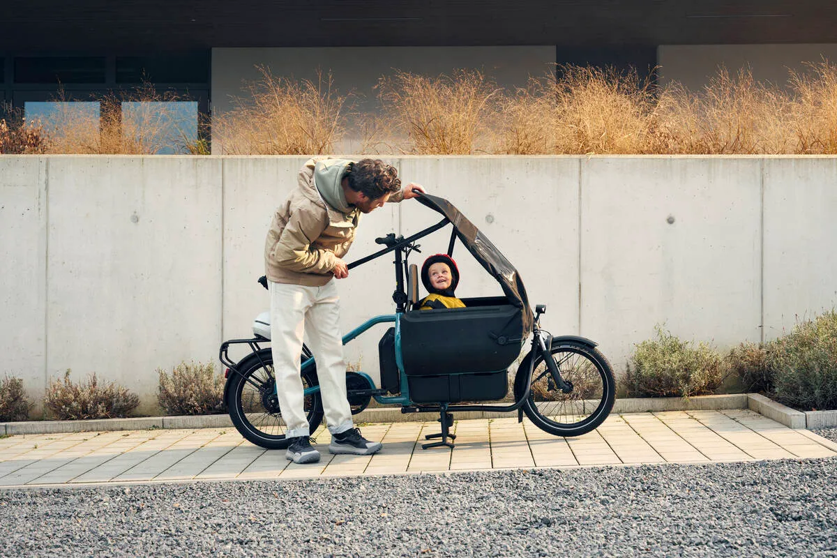 A parent with a child in an aqua Riese & Müller Carrie e-cargo bike, against a concrete wall backdrop