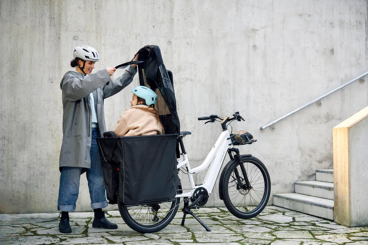 Woman in grey coat loading items into pearl white Multicharger2’s rear carrier outside modern building.