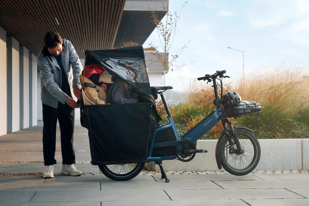 A person loads cargo into a blue Riese & Müller Multitinker e-bike with front carrier and weather protection.