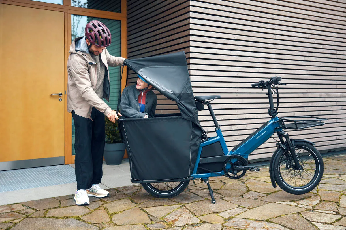 Cyclist with R&M Multitinker e-cargo bike shows covered passenger compartment outside modern home entrance.