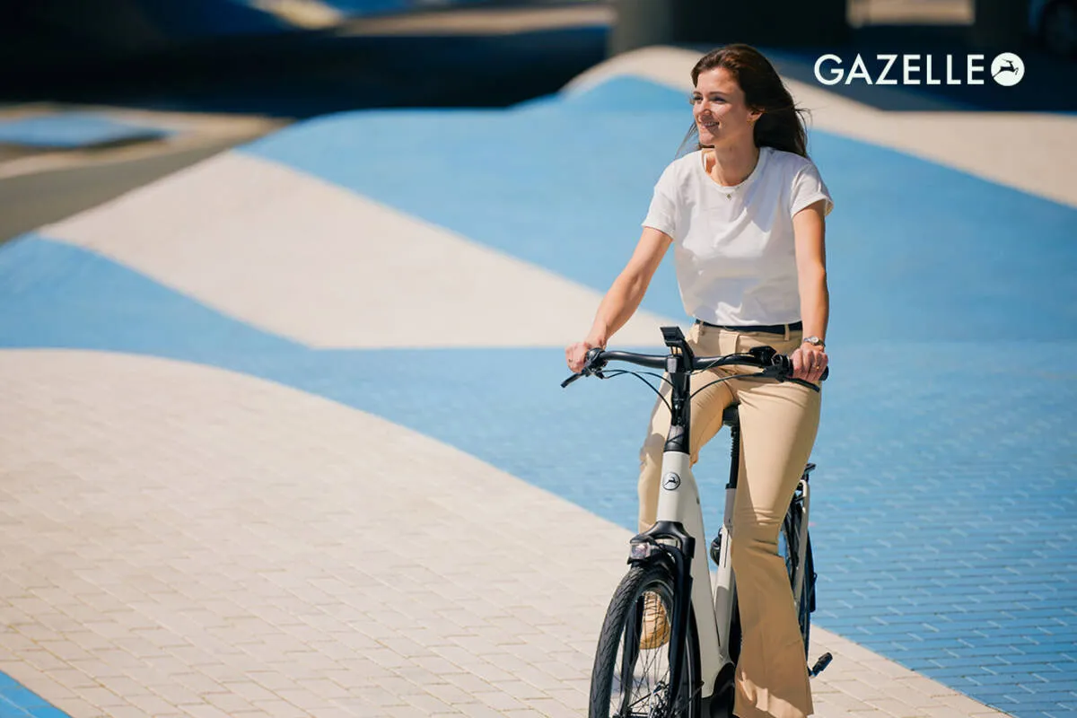 Woman in casual wear riding ivory white Gazelle Grenoble C5 e-bike on modern urban plaza.