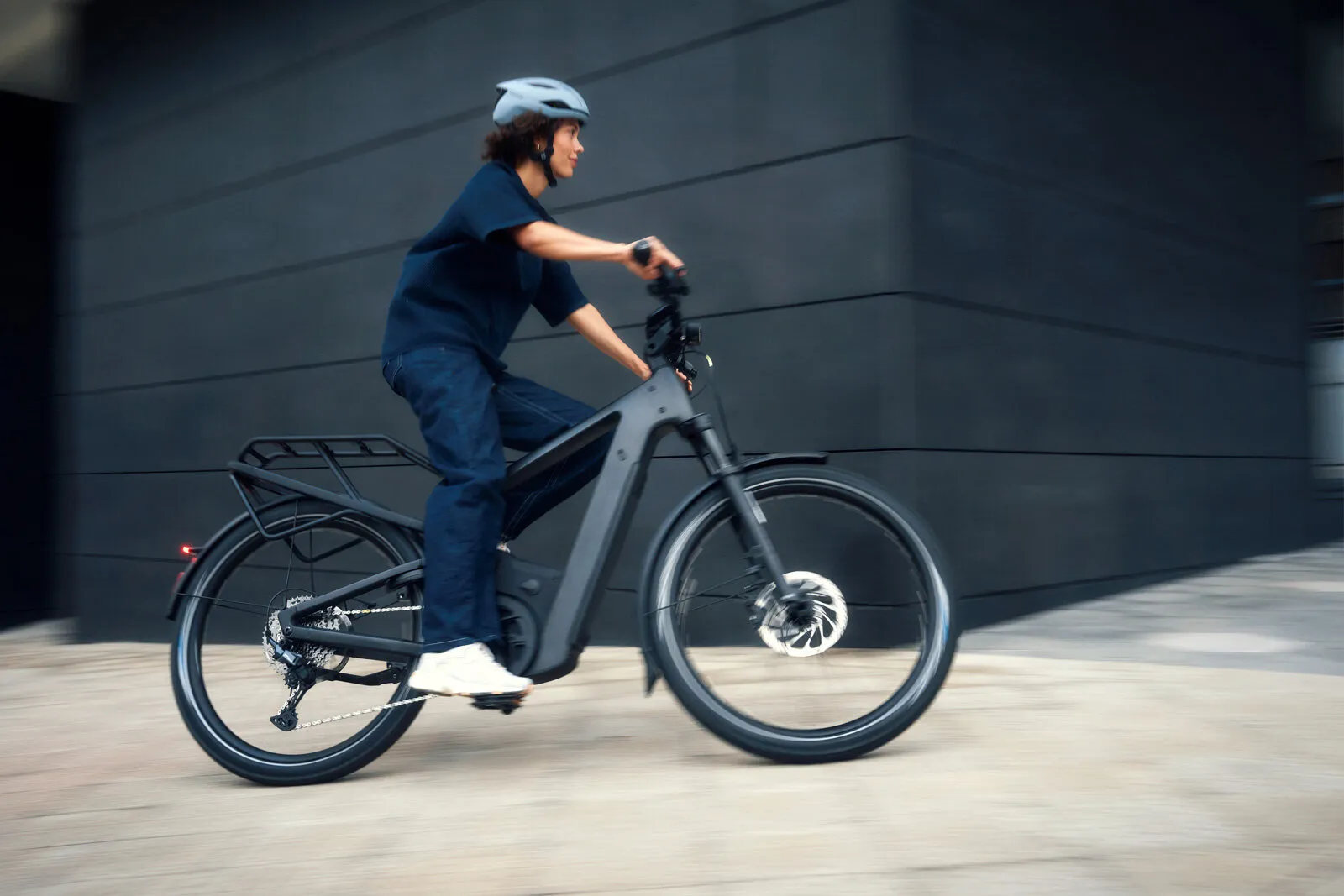 Woman riding Riese & Müller Delite5 in stone grey with rear rack, motion blur showing speed.