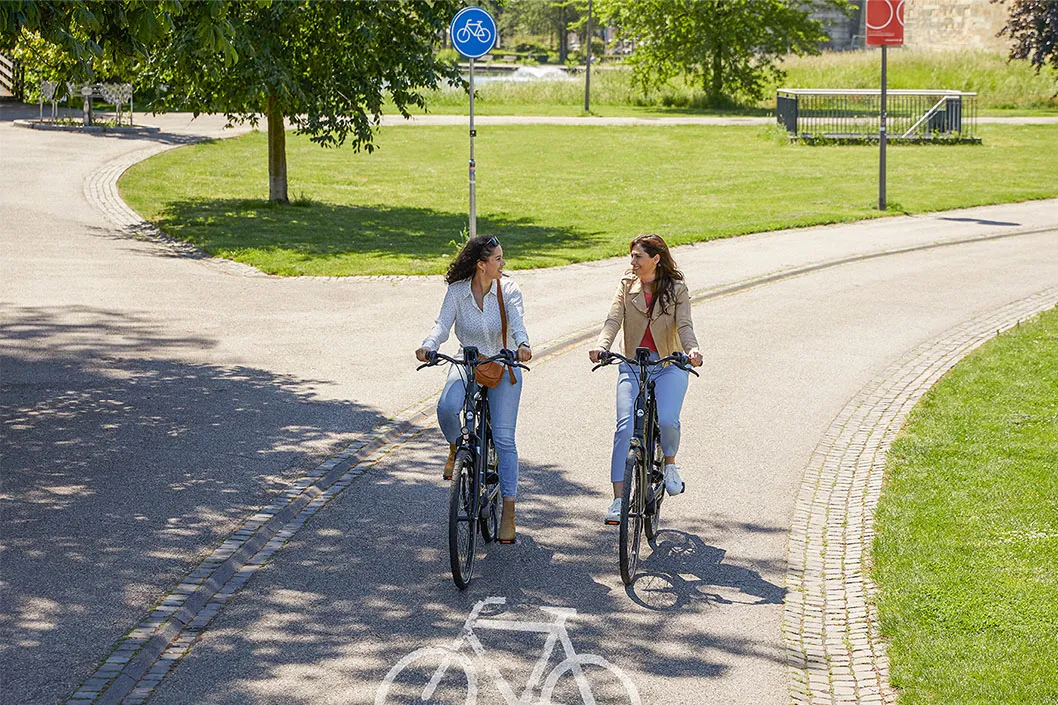 Two people riding their Gazelle e-bikes along a cycle lane through a park.