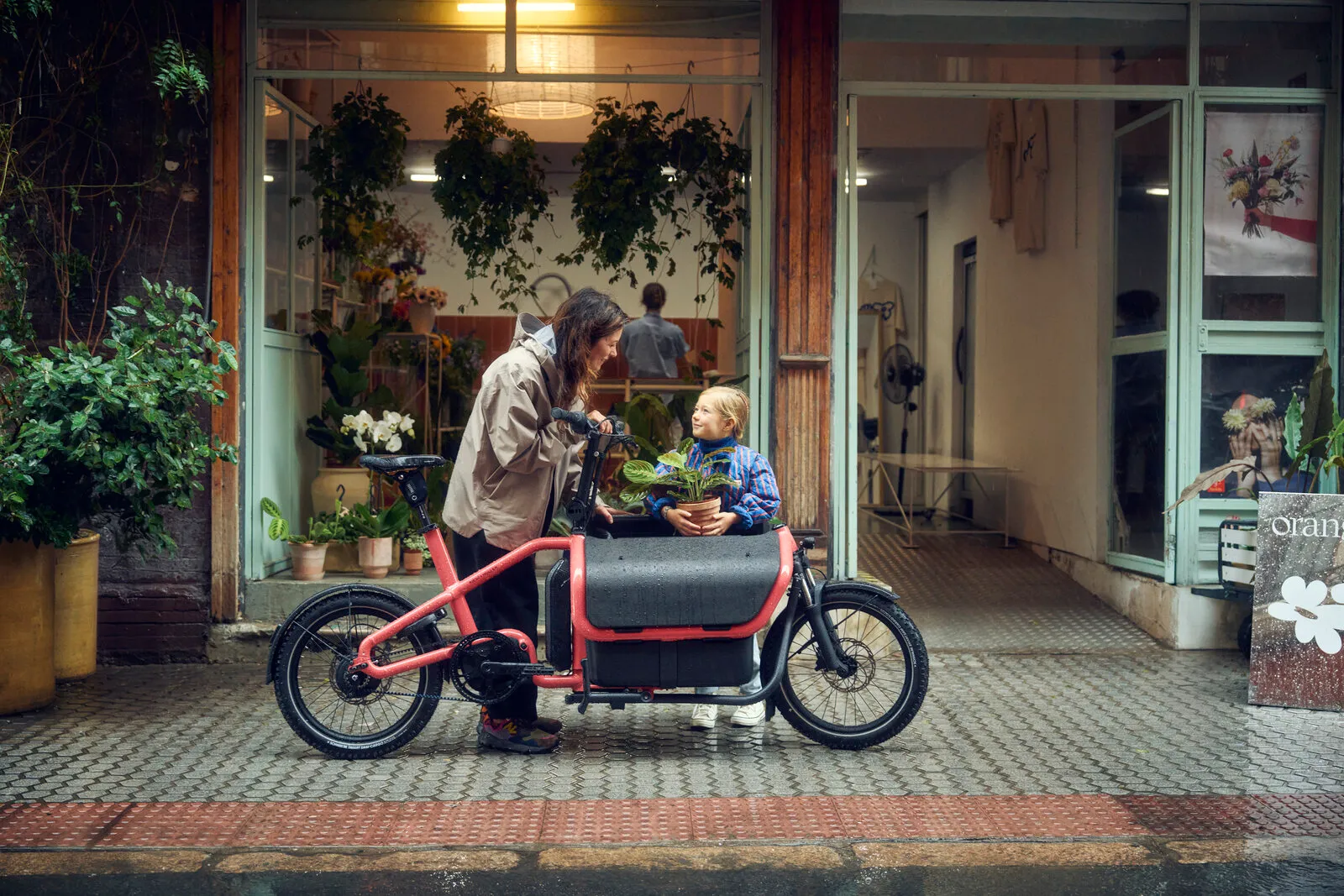 Riese & Müller Carrie2 vario in coral parked outside a flower shop, with a child sitting in the Flex Box holding a plant.