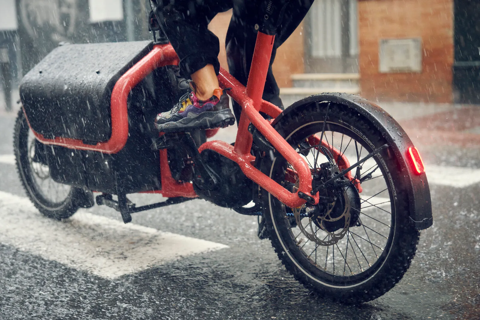 Close-up of the Carrie2 vario in coral being ridden in heavy rain, showing the rear wheel, disc brake and integrated tail light.