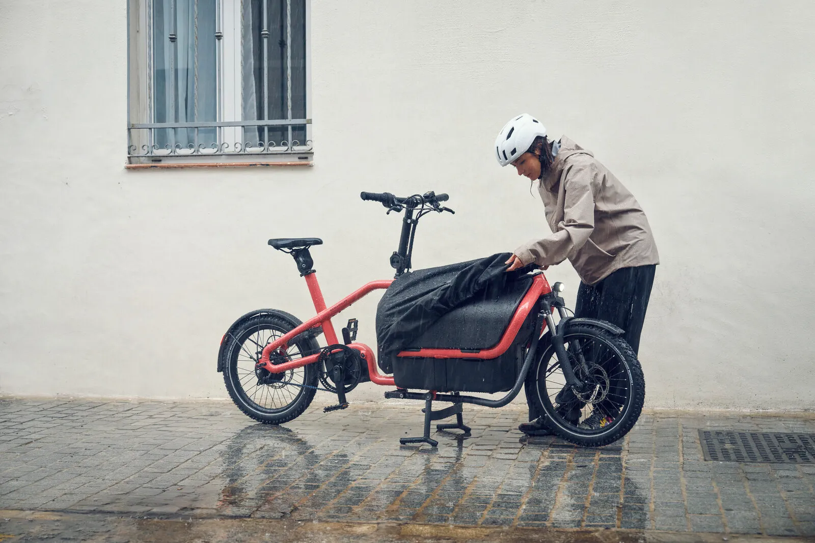 Rider fitting a rain cover over the Carrie2 vario Flex Box in coral, parked on a cobbled street in the rain.