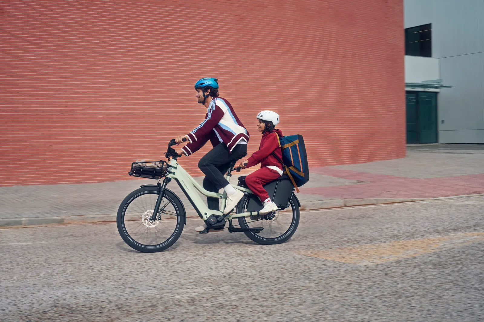 Rider and child on the Multicharger3 Mixte vario in desert/black, side profile passing a red building.