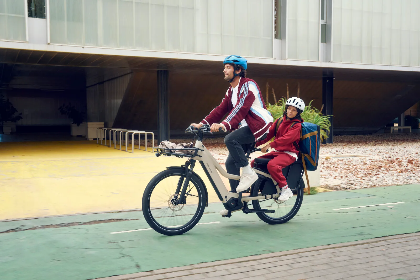 Rider cycling the Multicharger3 Mixte vario in desert/black with a child on the rear seat, front three-quarter view on a cycle lane.