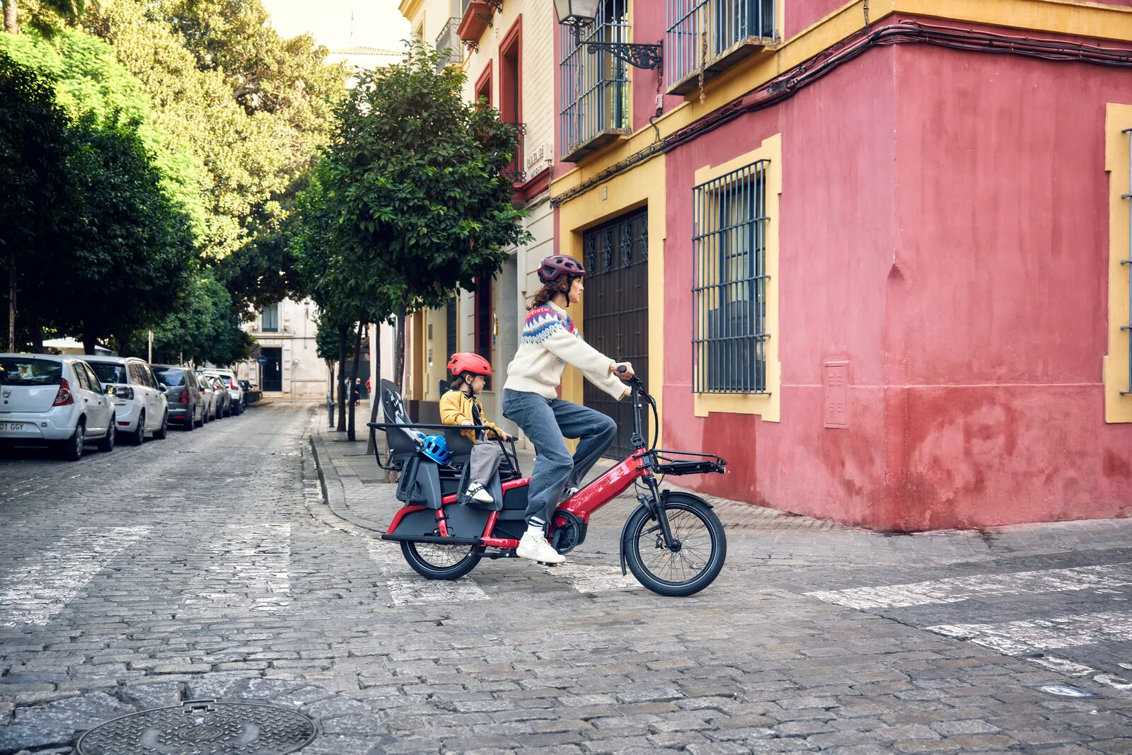Rider cycling the Multitinker2 vario in lava/black with a child on the rear seat, riding through a cobbled city street.