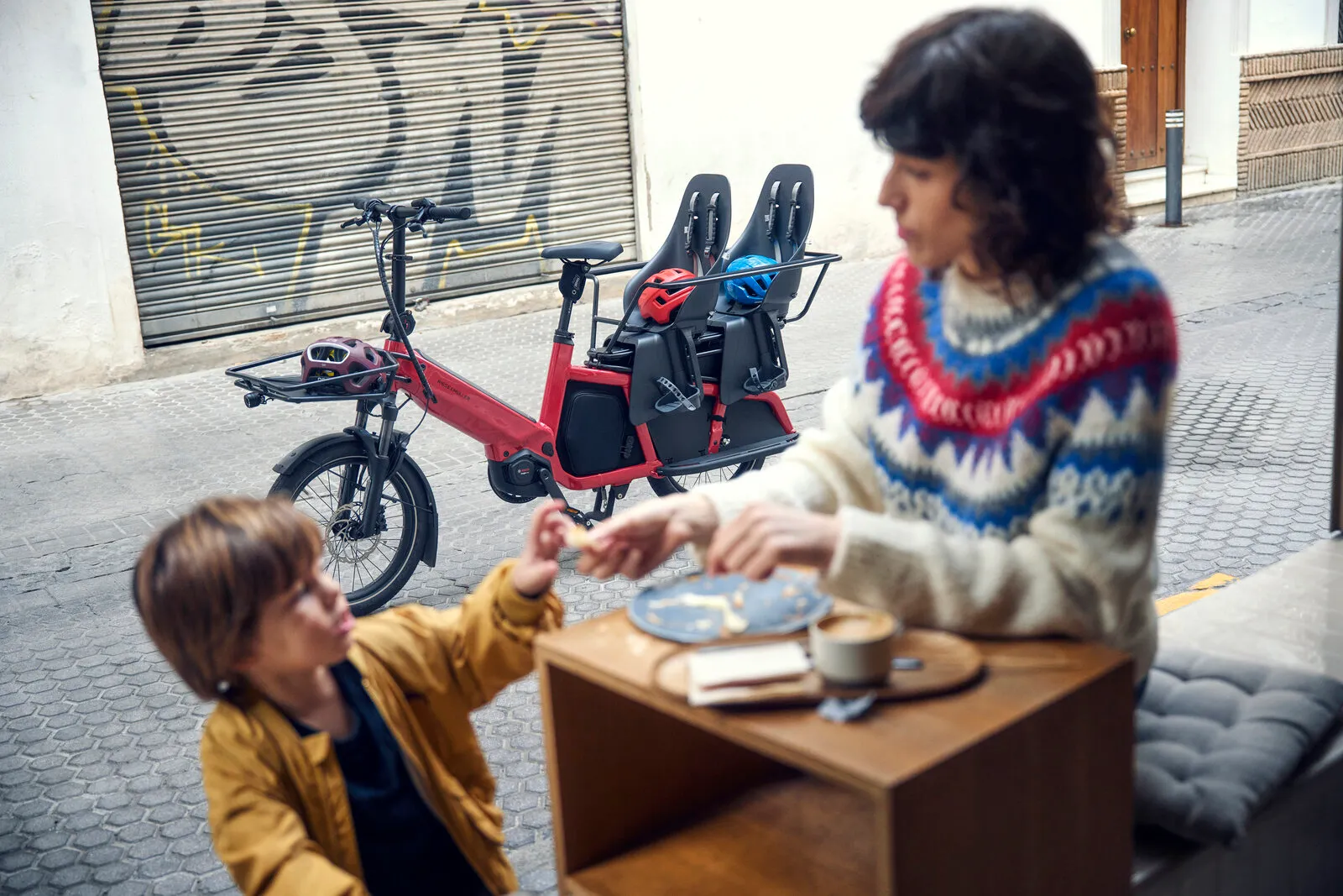 Multitinker2 vario in lava/black with child seats parked outside a café, with an adult and child sharing a snack at a table.