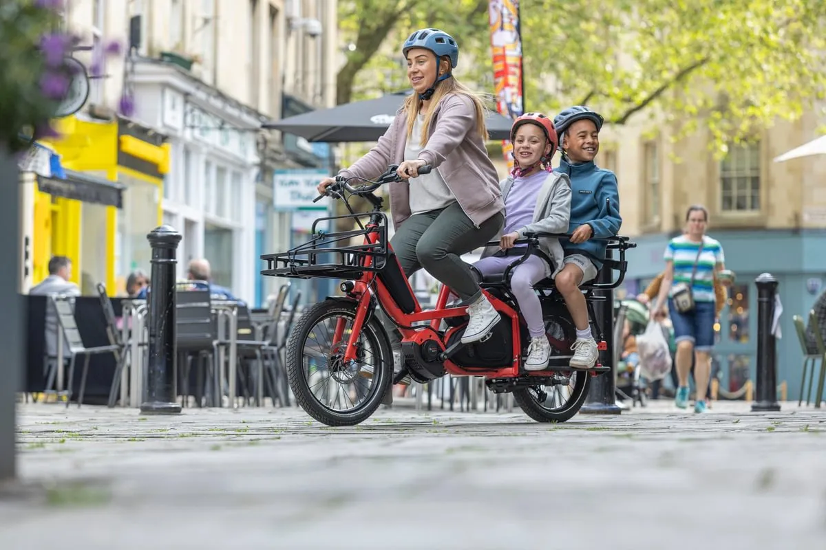 Woman riding red Tern Quick Haul Long e-bike with two children seated on rear platform through historic urban street with period buildings