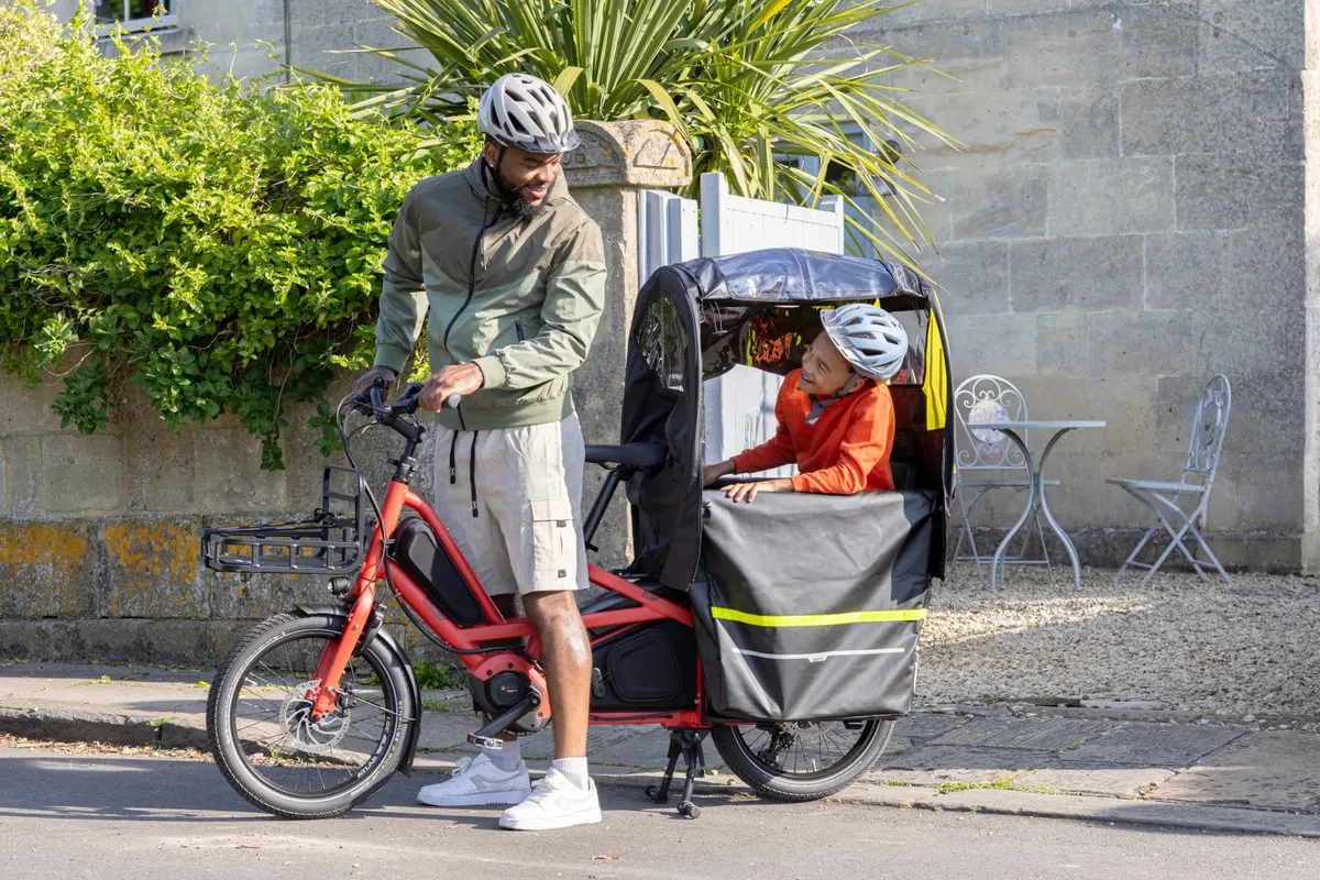 Man in cycling gear standing beside red Tern Quick Haul Long e-bike with child in attached bike trailer, urban setting with stone walls and outdoor café seating