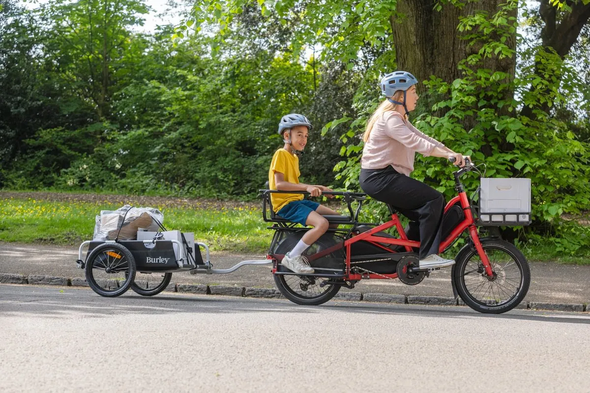 Woman cycling on red Tern Quick Haul Long e-bike with child passenger on rear platform, towing Burley trailer on tree-lined park path