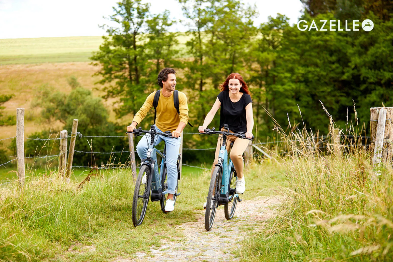 A couple riding Gazelle e-bikes side by side on a rural path, surrounded by summer meadow grass and trees.