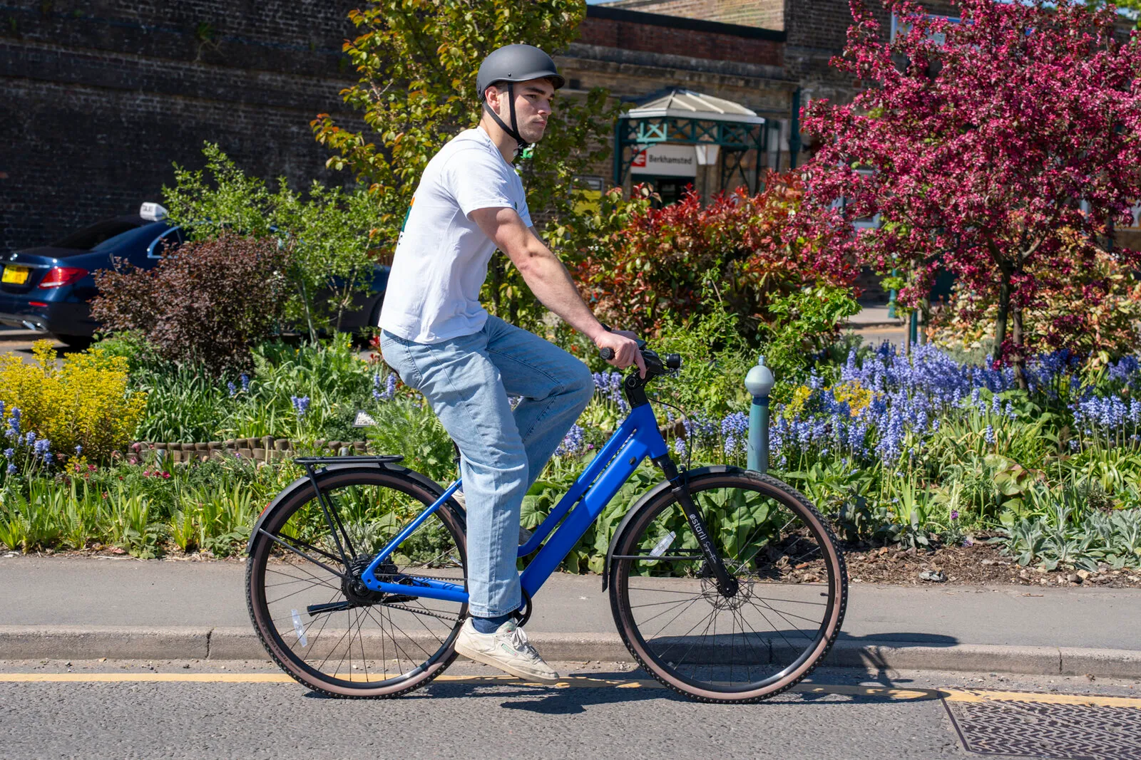 A man riding the Estarli e28.X step-through in cobalt blue past flower beds outside Berkhamsted station.