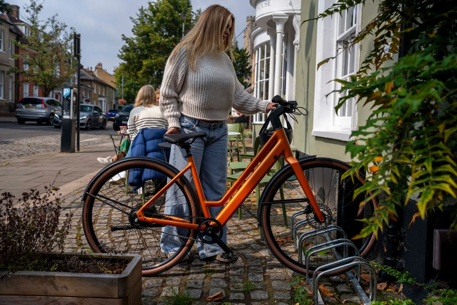 A woman parking the Estarli e28.X step-through in bronze at a cycle rack outside a pub.