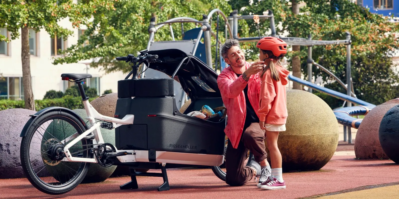 A father helping his daughter put on a helmet next to a Riese & Müller Packster cargo bike at a playground
