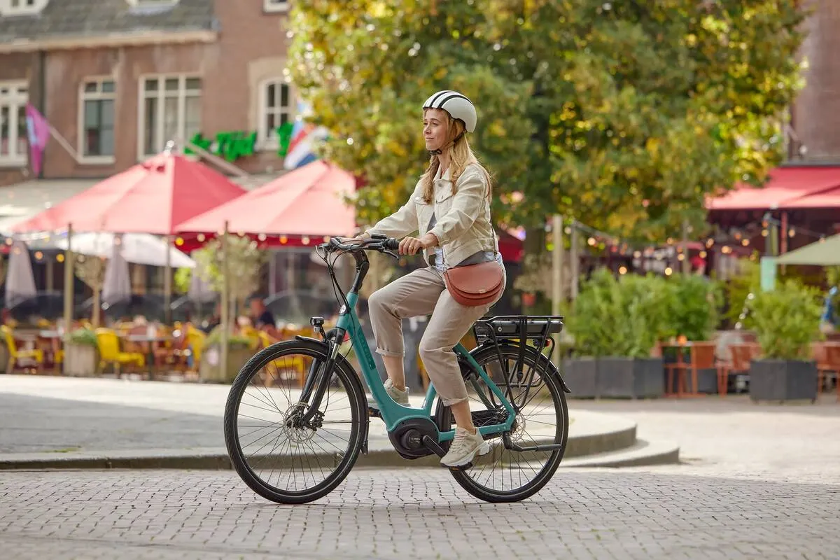 Woman riding a thyme green Gazelle Paris C7+ e-bike on a cobbled street, with cafe terraces and pink umbrellas in the background.