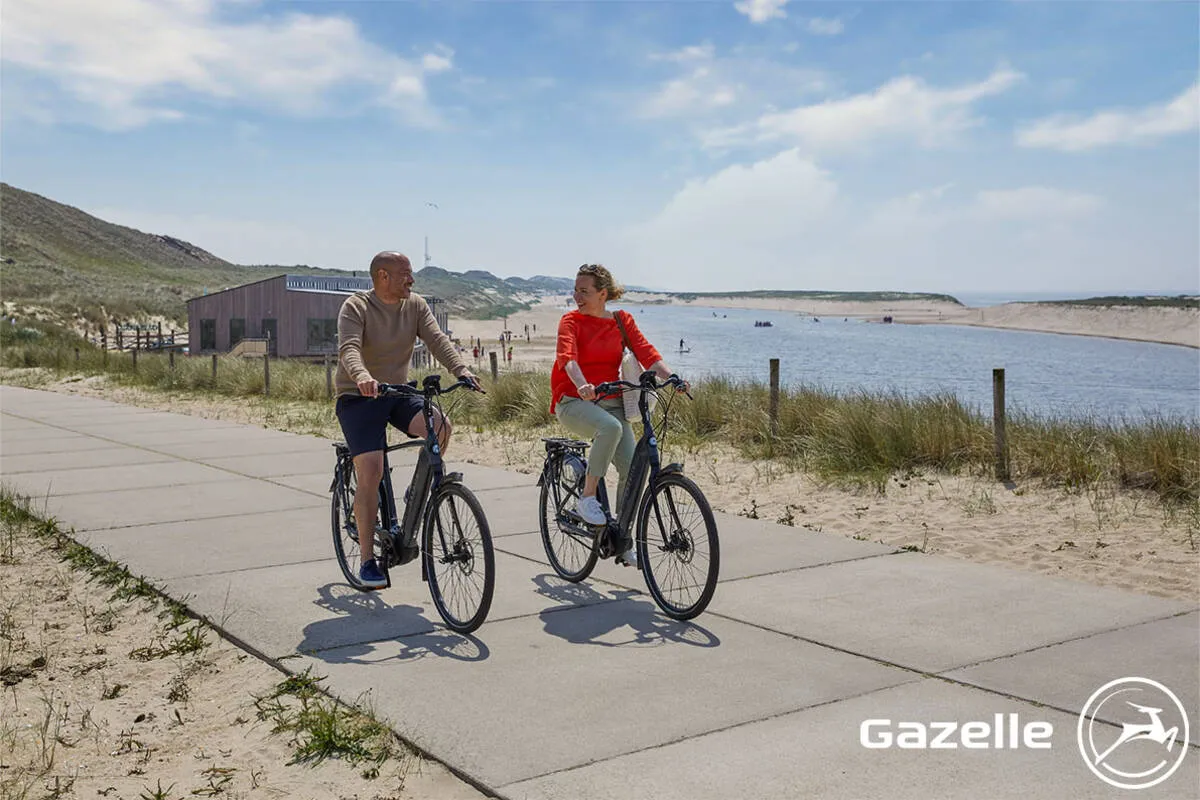 A couple riding Gazelle Grenoble bikes along a beachfront path.