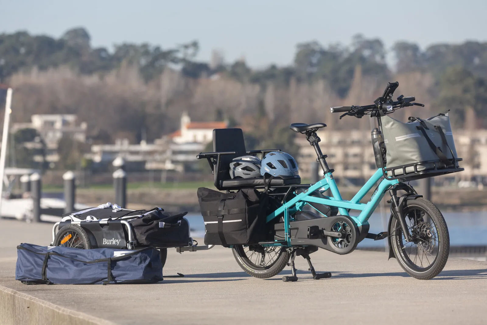 A beetle blue Tern GSD loaded with panniers, a child seat, a front rack bag and a Burley trailer at a marina.