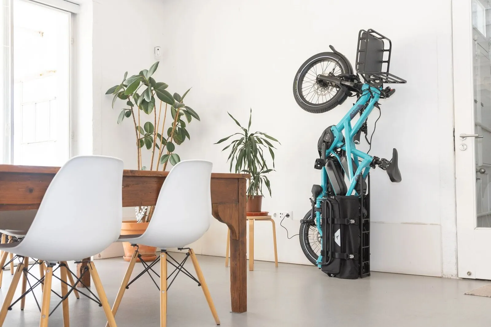 A beetle blue Tern GSD parked vertically on its rear rack against a wall, next to a dining table.
