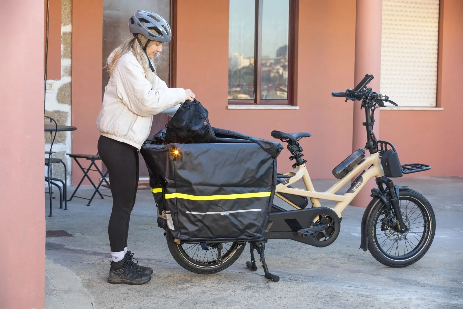 A woman loading a large rear cargo bag on a satin beige Tern GSD outside a café.