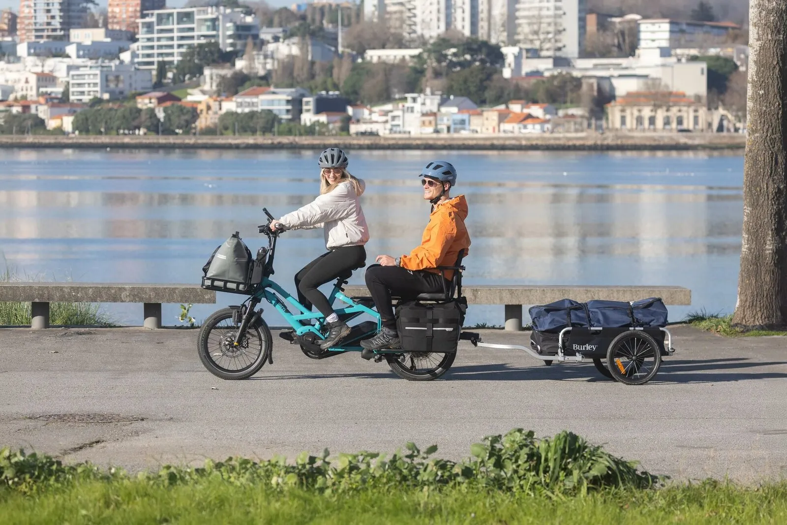 A woman riding a beetle blue Tern GSD with an adult passenger on the back while towing a Burley trailer.