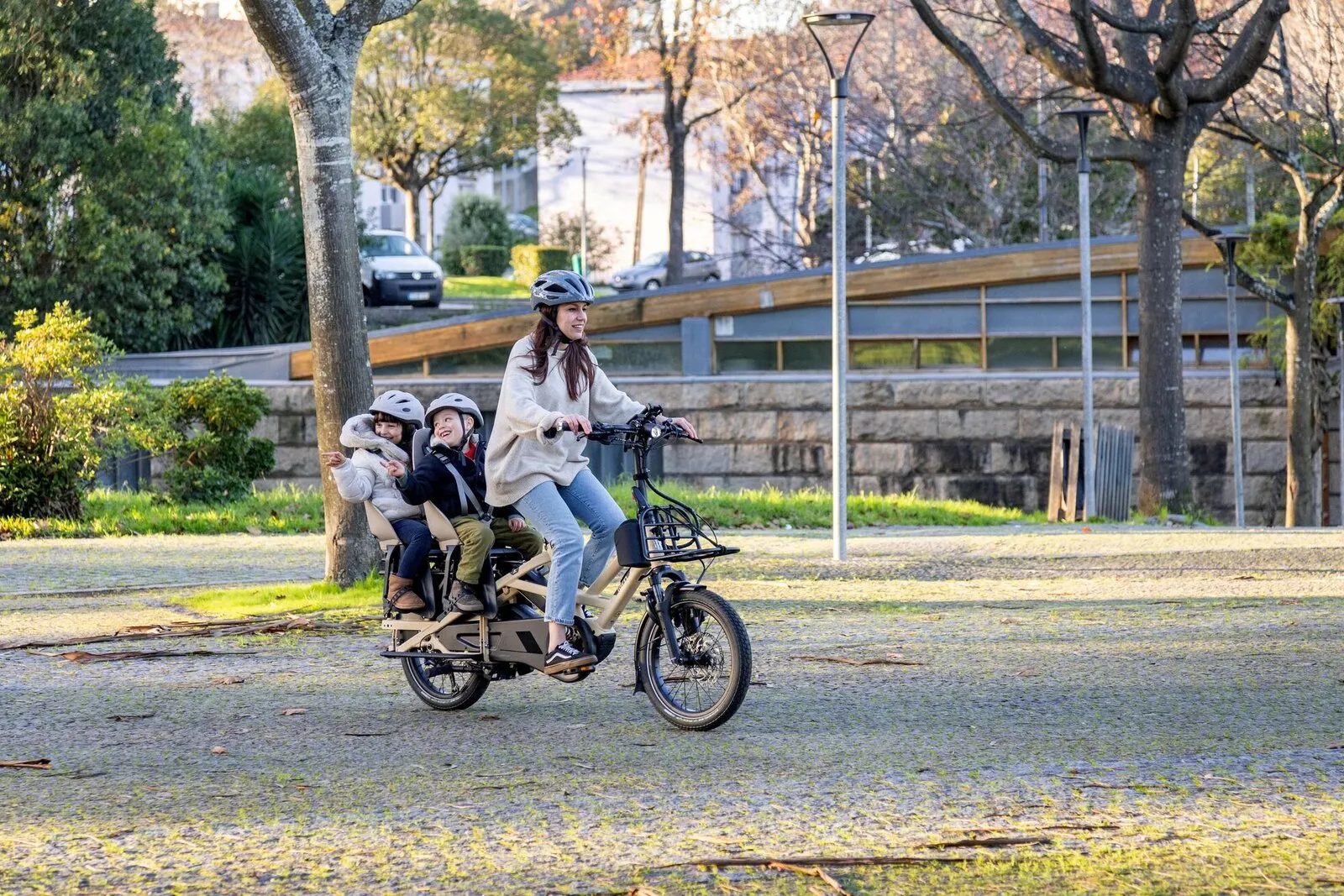 A woman riding a satin beige Tern GSD through a park with two children seated on the back.