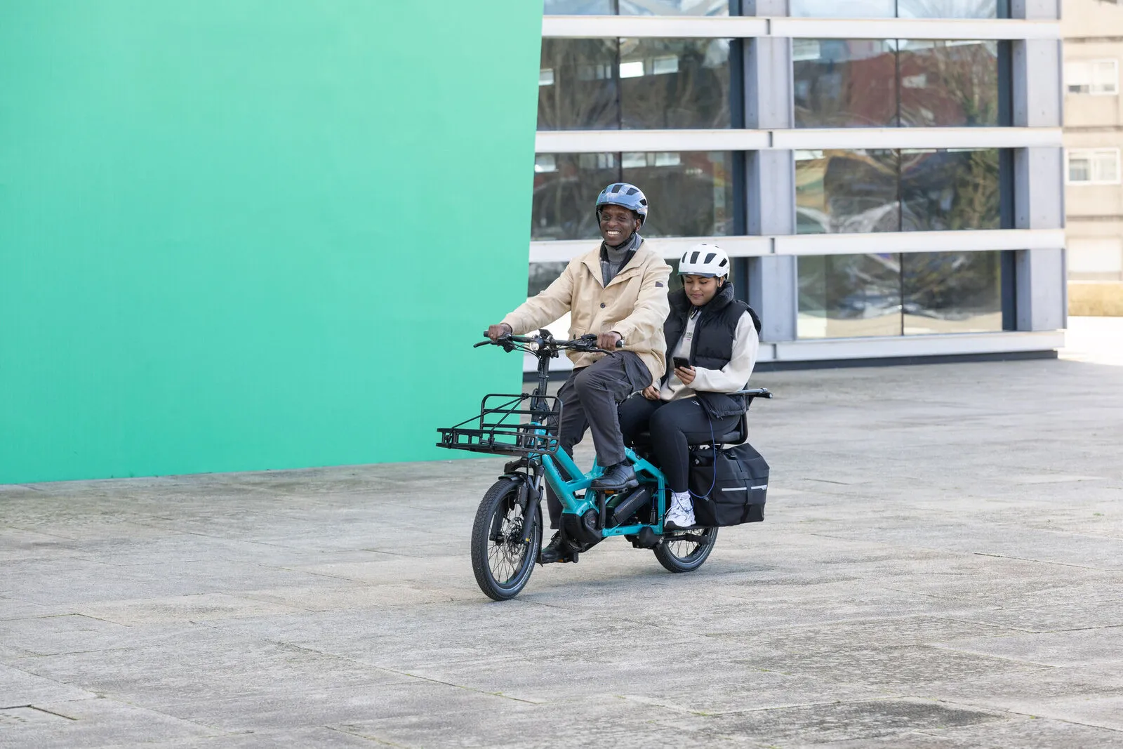 A man riding a beetle blue Tern GSD with his daughter as passenger, in front of a turquoise wall.