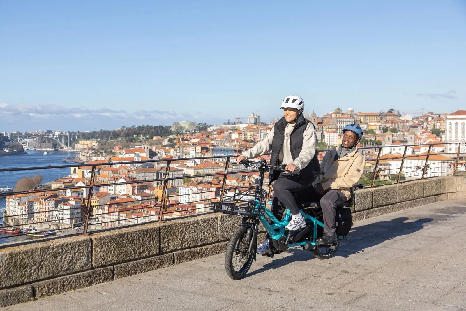 Two adults riding a beetle blue Tern GSD along a riverside promenade, one as passenger on the rear deck.