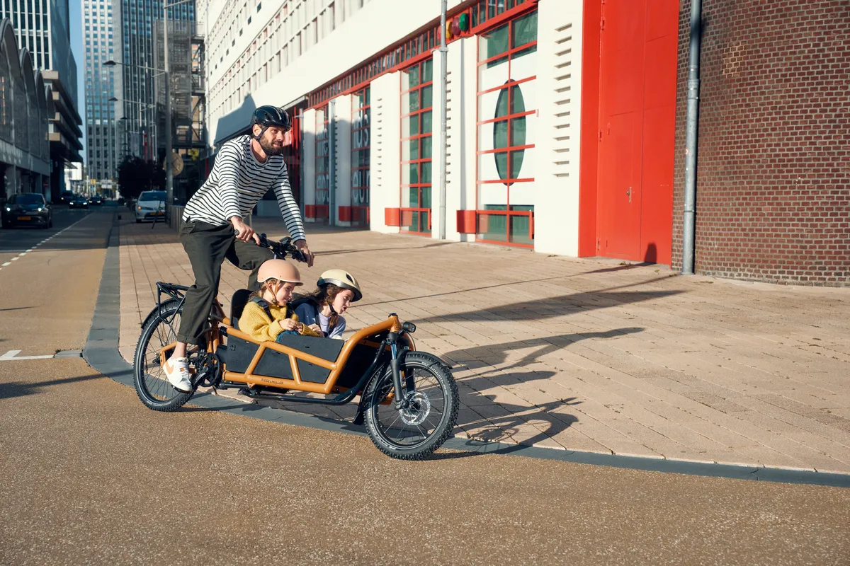 A man riding a Riese & Müller Load4 75 in peanut matt on a cycle lane carrying two young children.