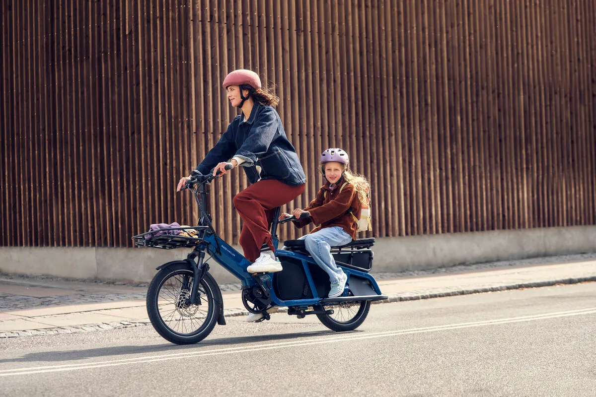 A lady riding a petrol blue Riese &amp; M&uuml;ller Multitinker family bike with a young girl on the back.
