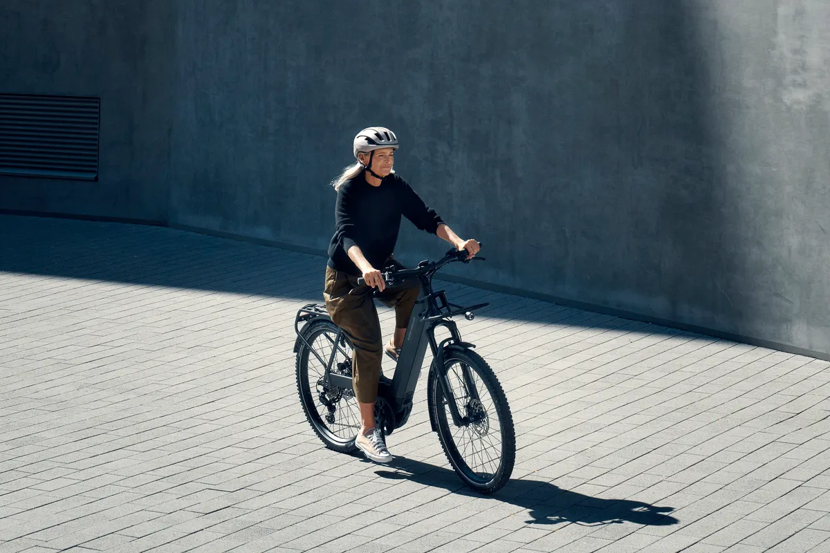 A lady riding a dark grey matt Nevo4 e-bike along a paved street scene.