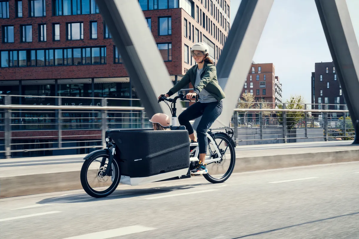 A lady riding a cargo bike on a cycle lane across a bridge. The bike is a Riese & Müller Load4 60 in a greenish grey colour the manufacturer calls tundra grey matt.