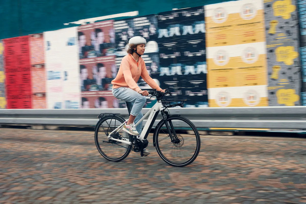 A lady riding a white Roadster e-bike along a paved street scene.