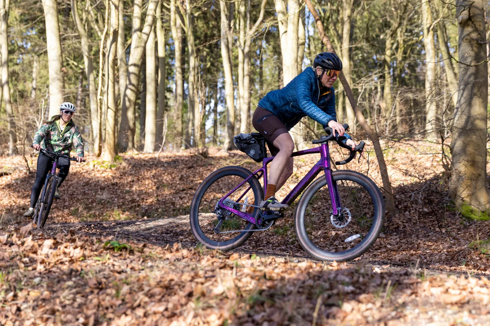 A woman riding a cosmic purple G700 down a woodland trail, with a second rider behind.