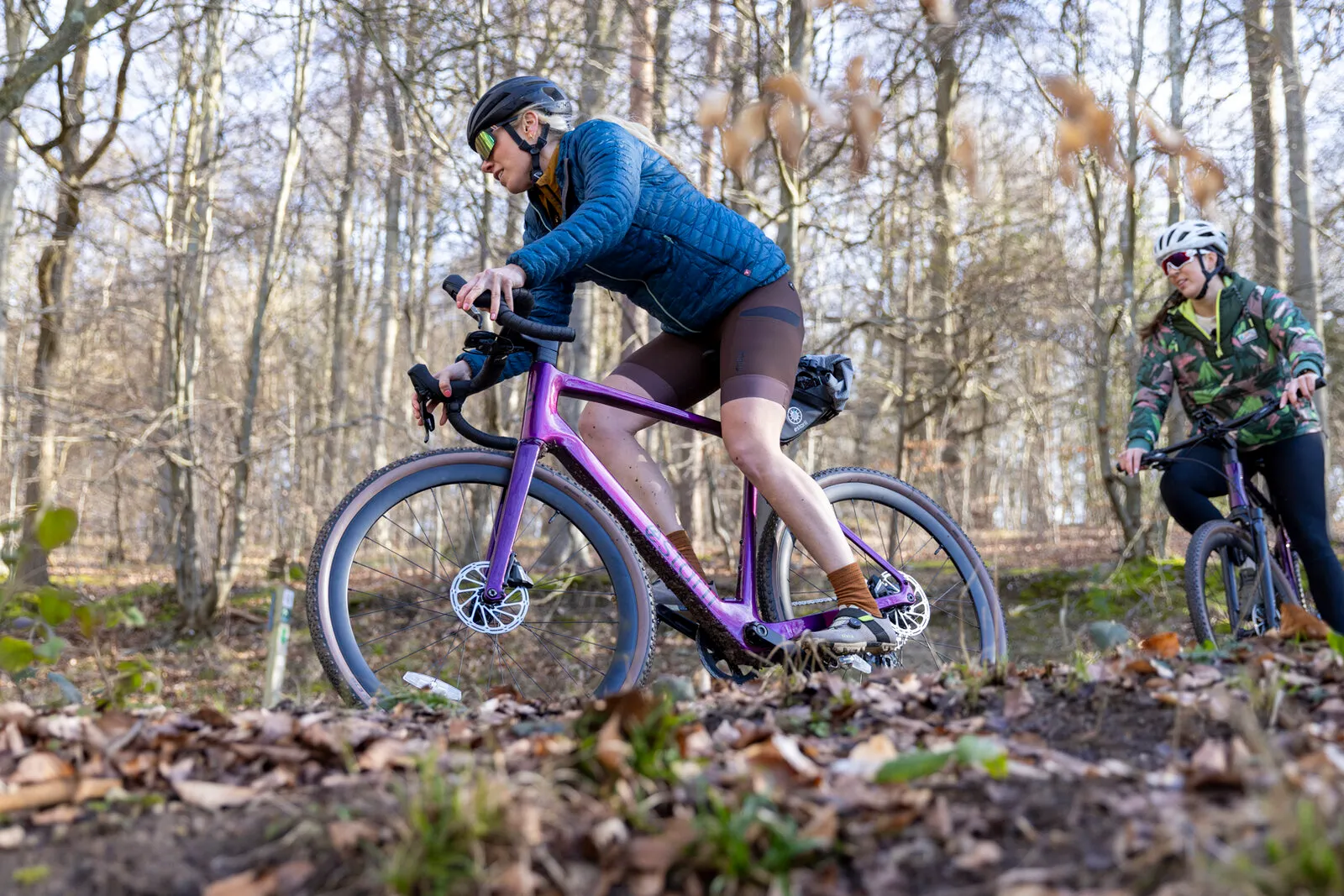 A woman riding a cosmic purple Estarli G700 up a woodland trail, with a second rider behind.